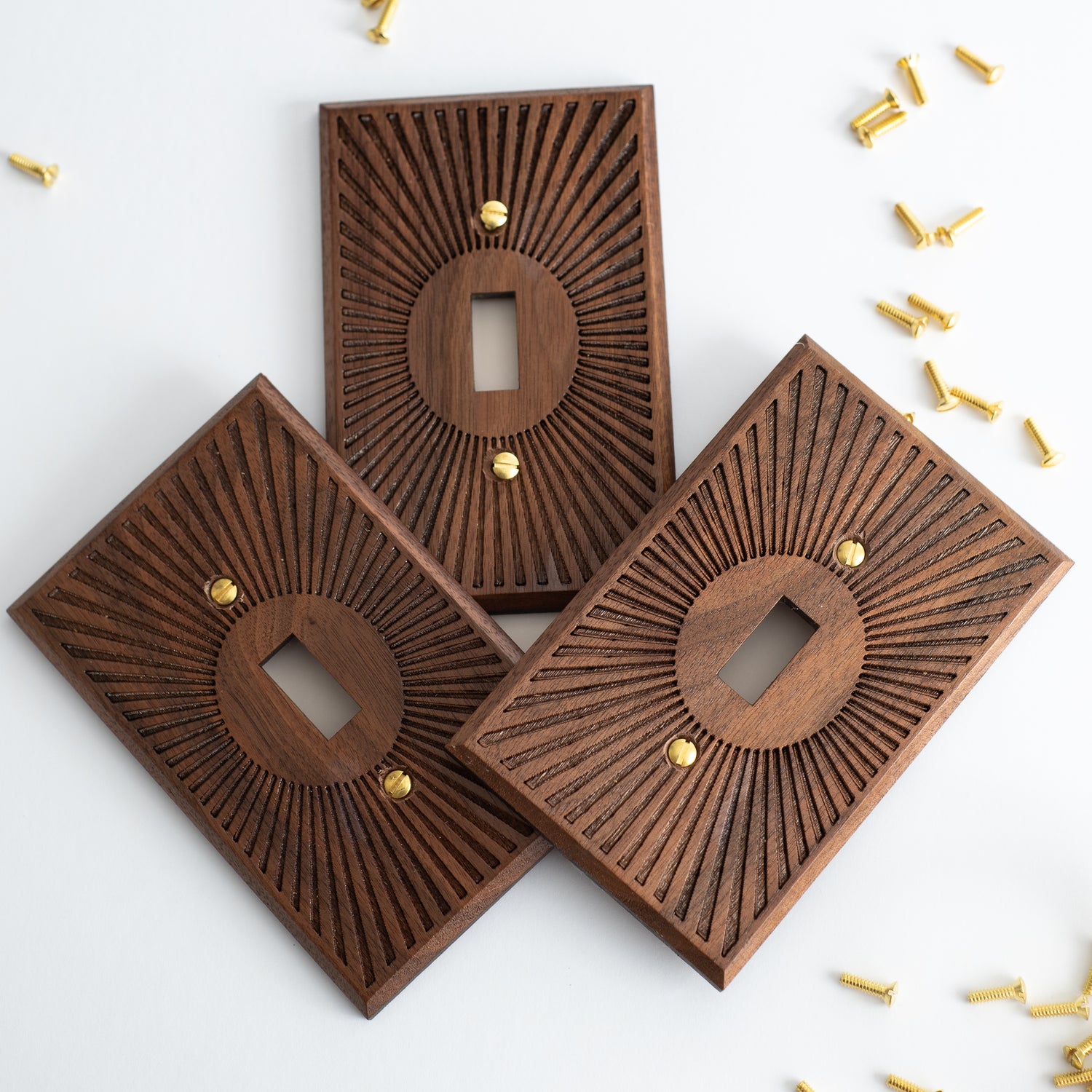 3 toggle switch walnut light switch plates on a white background with brass screws surrounding them on all sides. The engraving on the surface of the light switch plates resembles a sunburst  